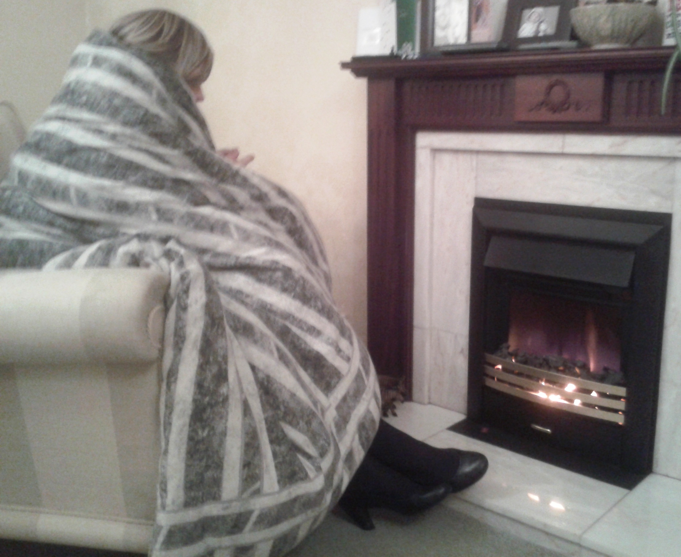 Elderly lady in front of a fire with a blanket around her keeping warm